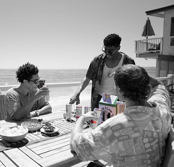 Four people sitting around a table by the beach, enjoying drinks and snacks.